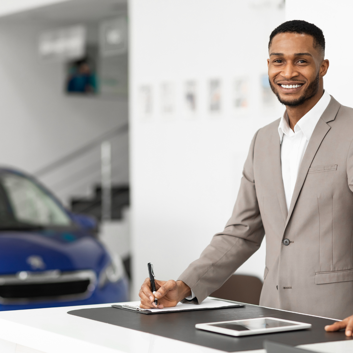 A car salesperson smiling with his iPad and his notes while writing on his notepad.