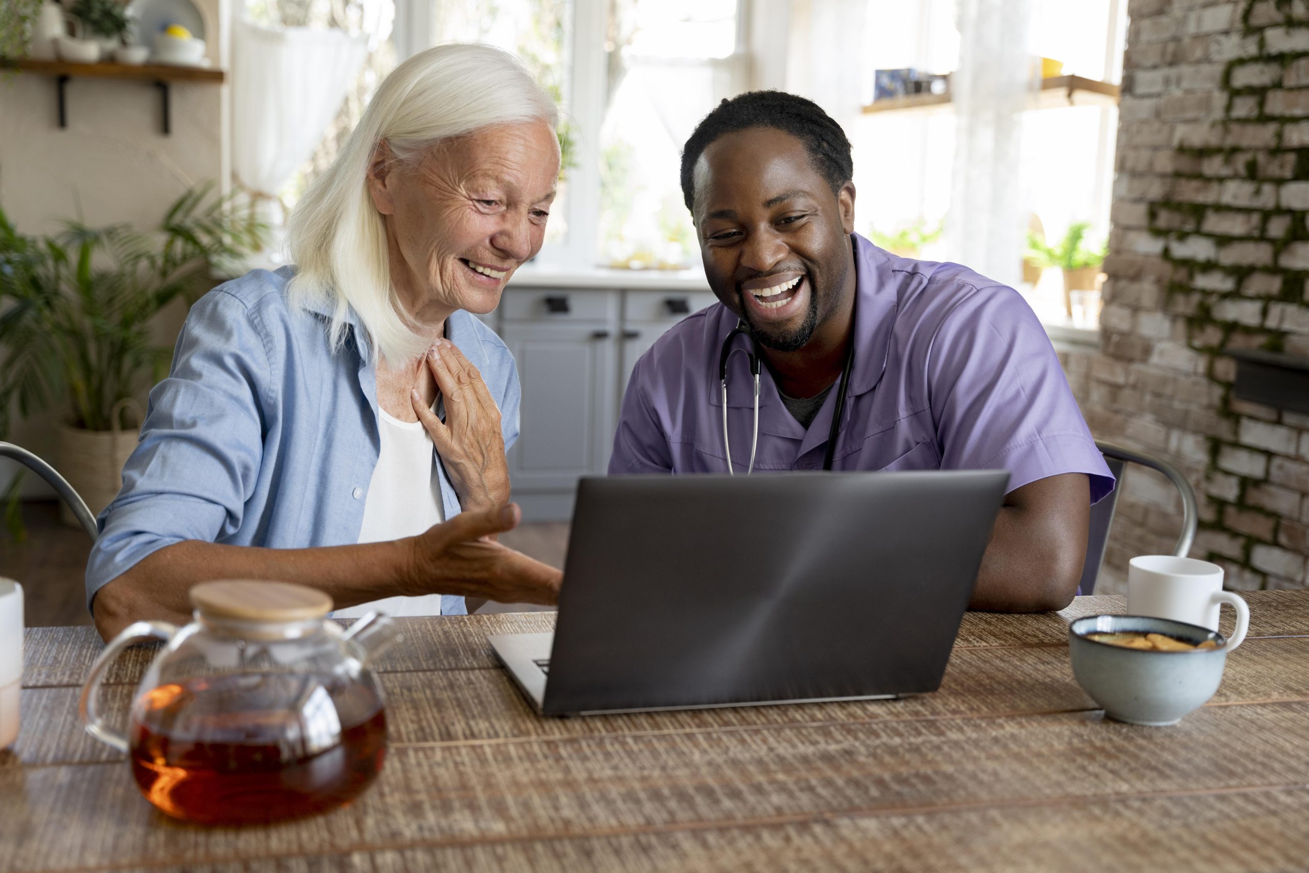 A smiley, friendly social worker in their scrubs is looking at a laptop screen while sitting at the breakfast table next to a senior person who is also smiling at the laptop screen.