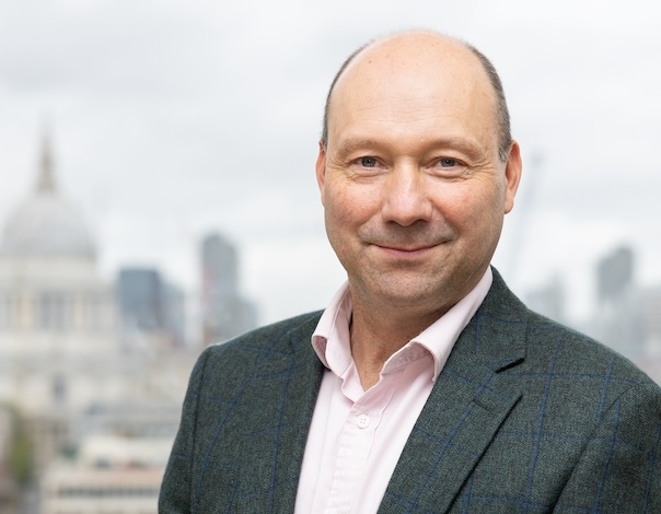 An image of Jolyon Parsons Ciptex's Strategic Director. Jolyon is smiling while wearing a light pink shirt with a slate grey blazer over the top of his shirt, with a blurred out backdrop of London city centre.